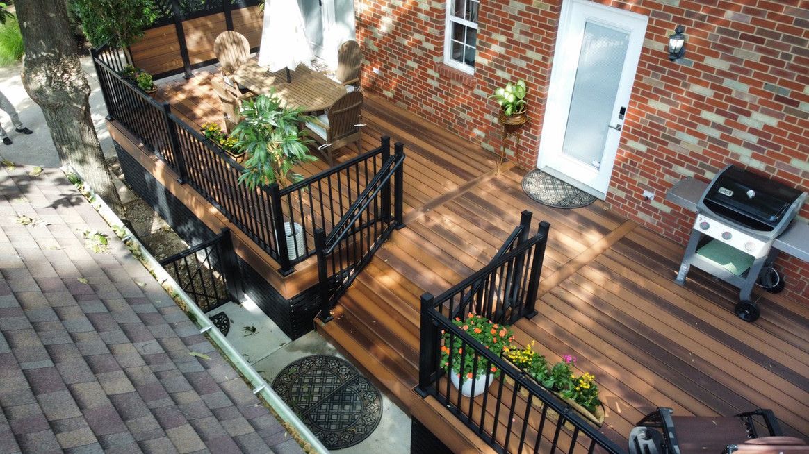 Elevated wooden deck with black railings and a brick house. Includes a grill and potted flowers.