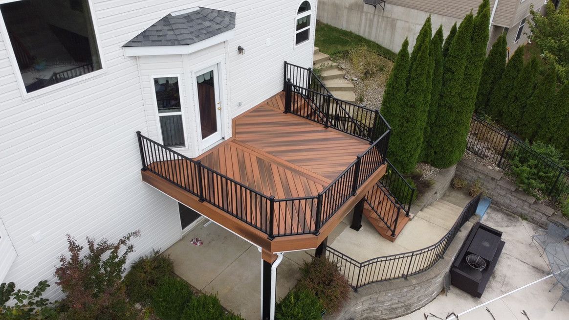 Deck with dark wooden planks, black railing, and stairs. Adjacent to a white house and greenery.