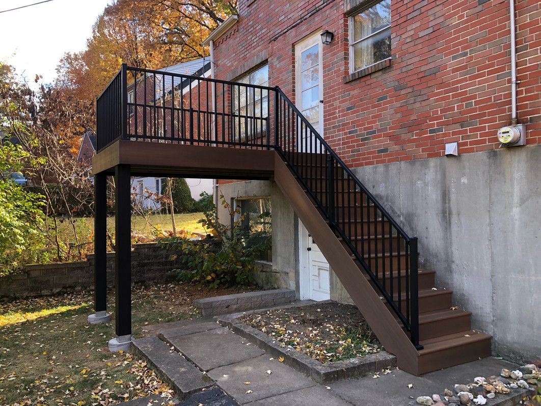 Deck and stairs attached to brick building, black railings, brown deck, and stairs, surrounded by a yard.