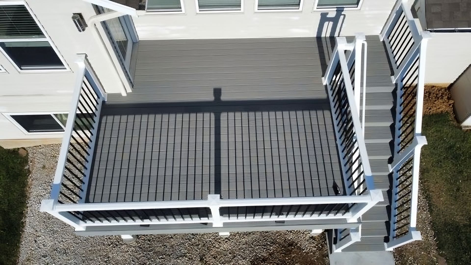Aerial view of a gray wooden deck with black accents and white railings, connected to a house with grass surrounding it.