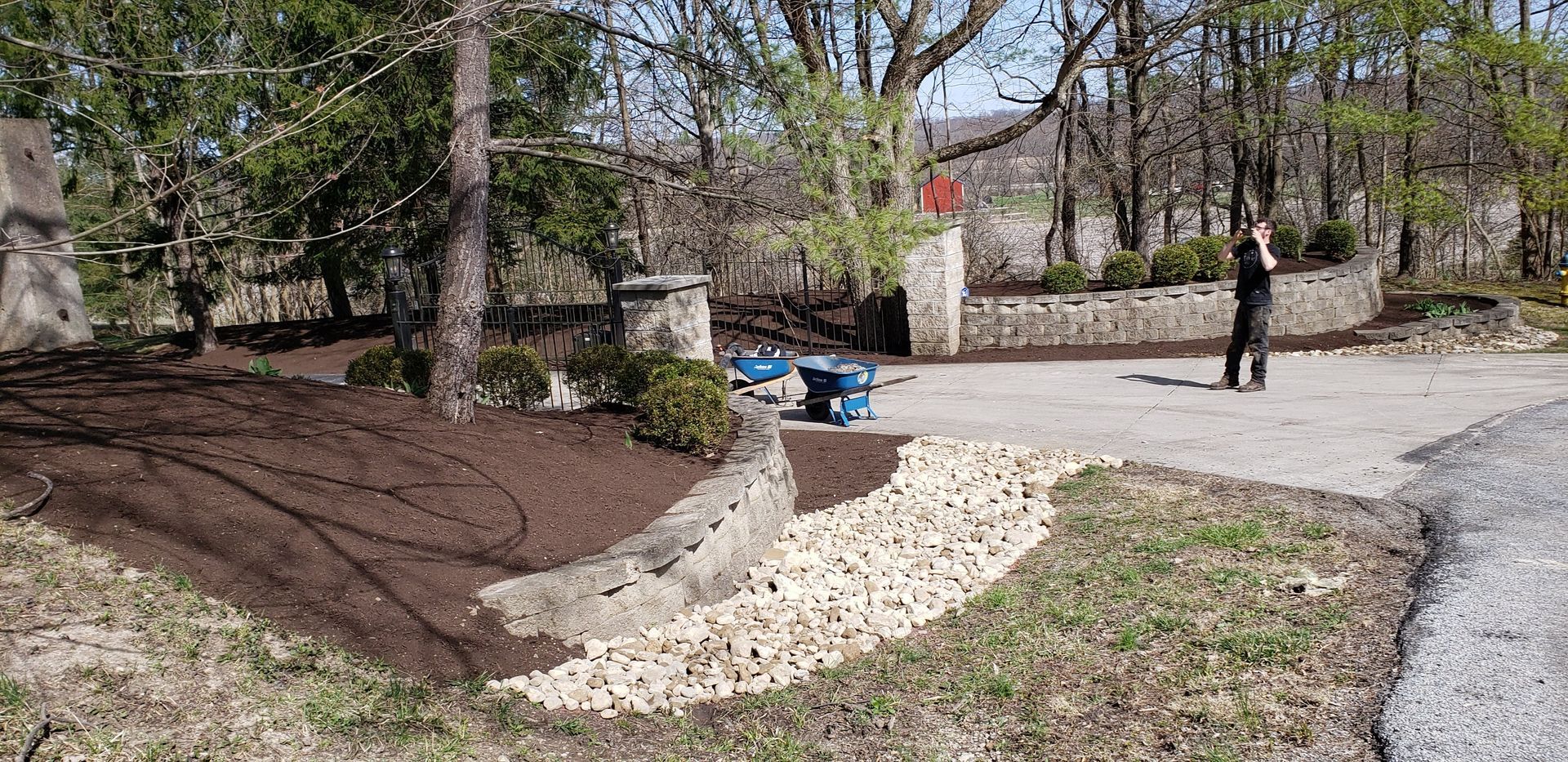 A person stands on a paved driveway with landscaping in front of a stone wall and trees.