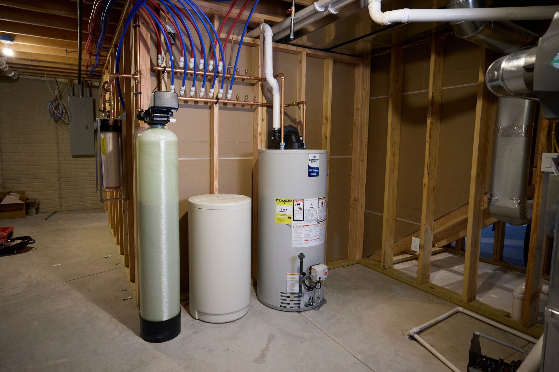 Basement with water heater, water softener, and plumbing. Wooden studs and concrete floor.