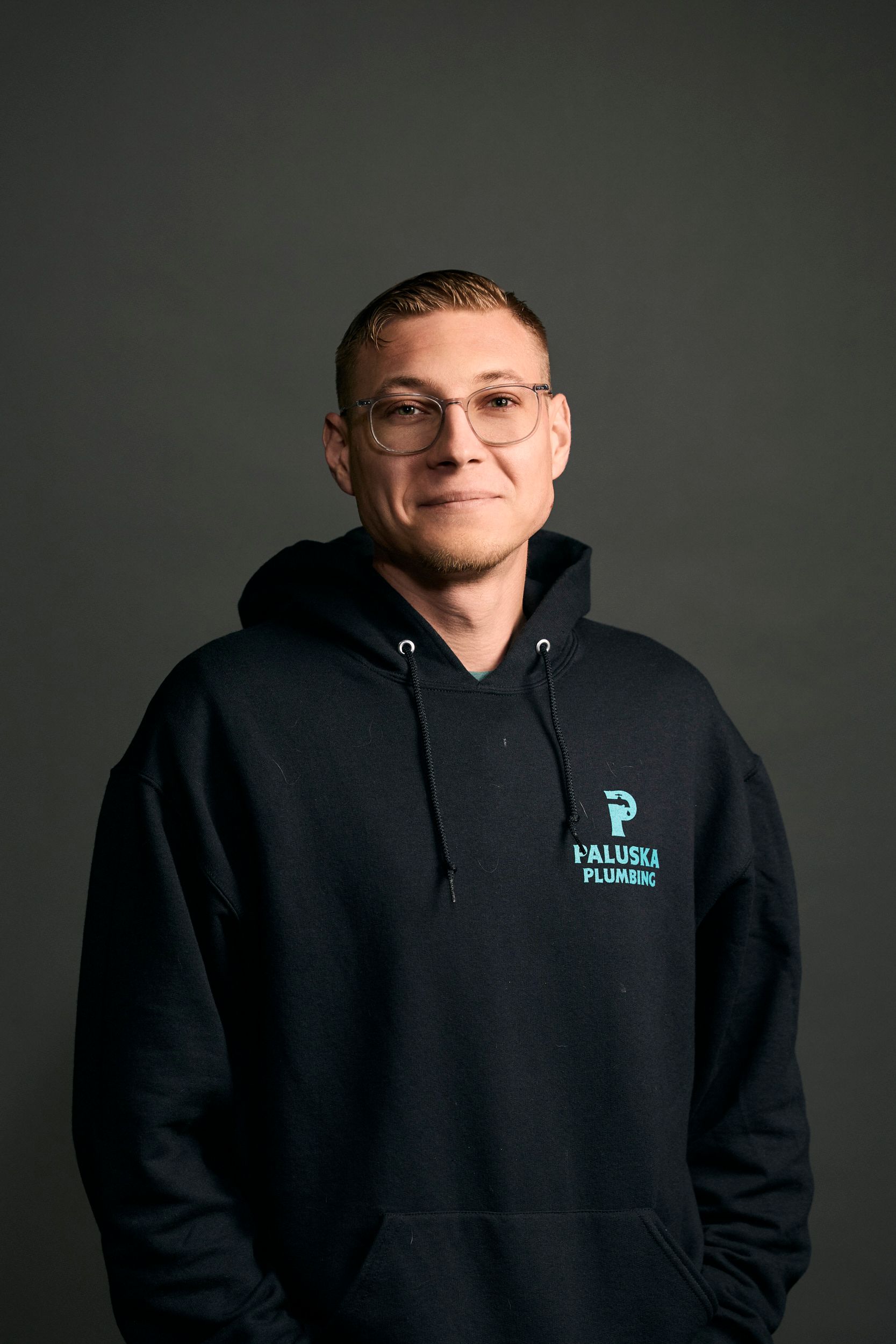 Man with glasses wearing a blue hoodie, smiling. Gray background.