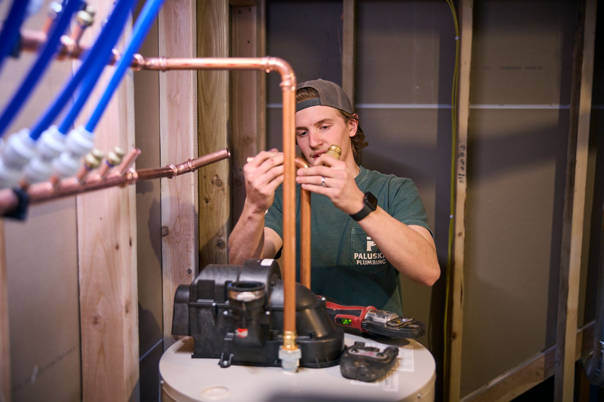 Man in cap and work clothes inspects pipes near the floor of a basement.