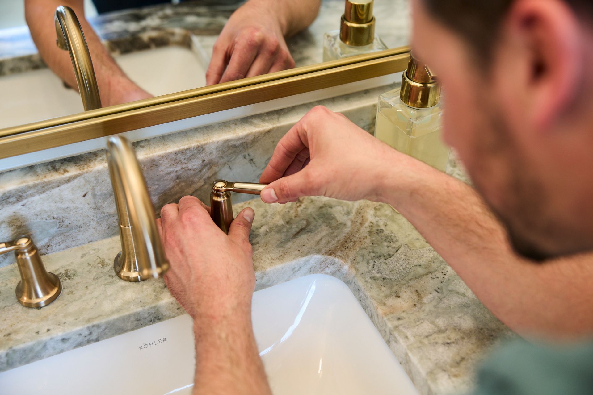 Man fixing a gold faucet in a bathroom with a marble countertop and mirror.