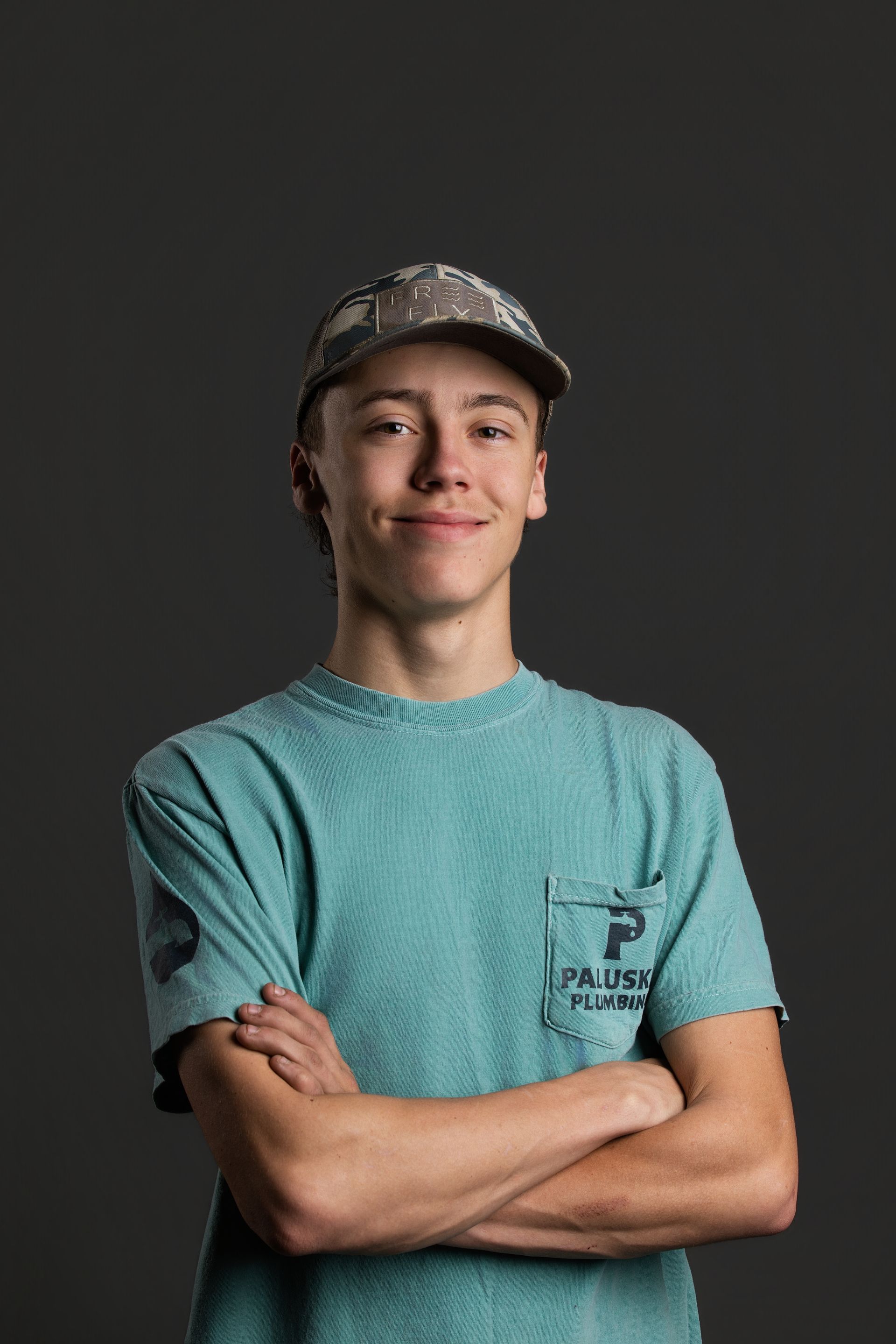 Person in a turquoise T-shirt and cap, standing with arms crossed against a dark background.