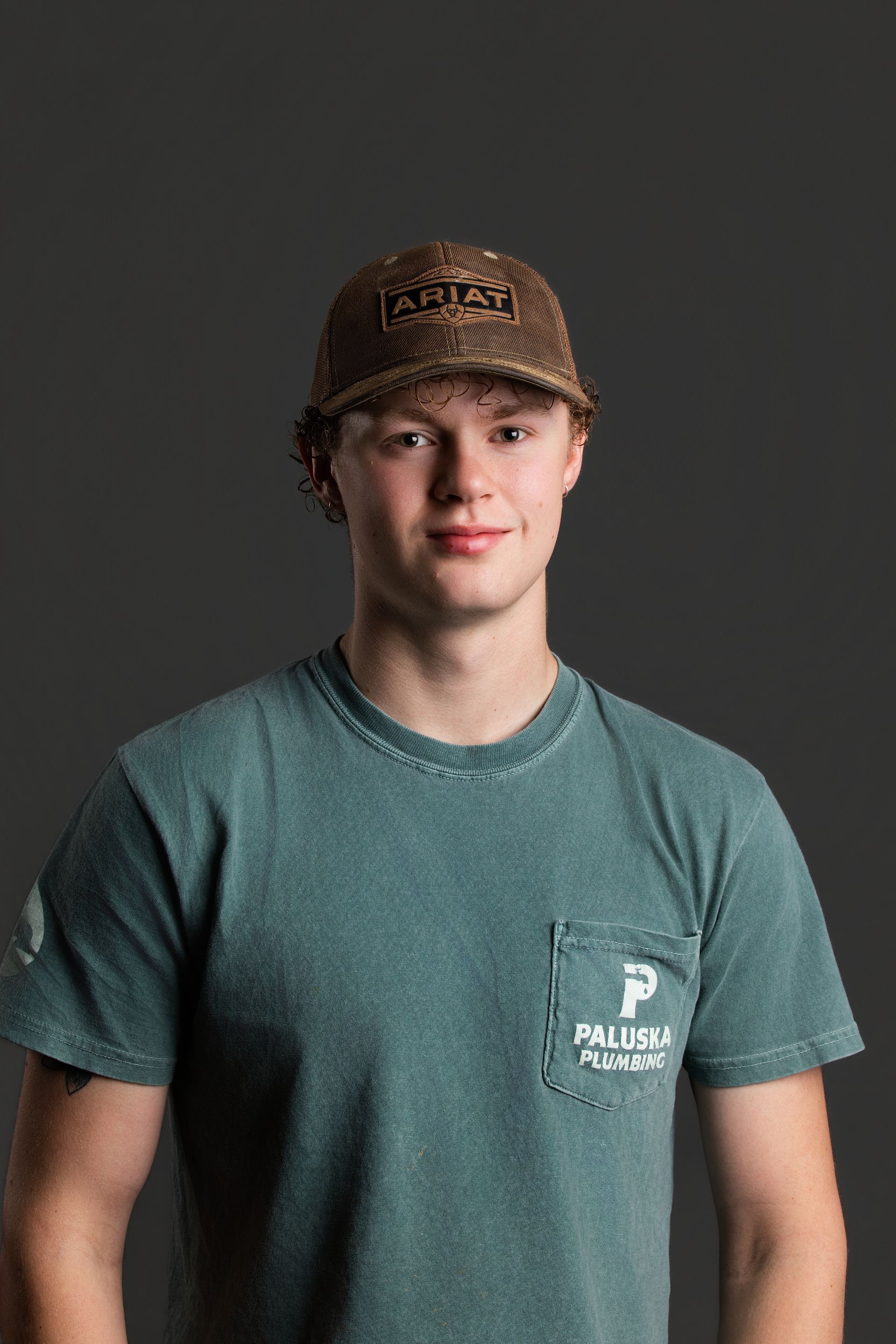 Person in a brown cap and teal T-shirt standing against a dark gray background.