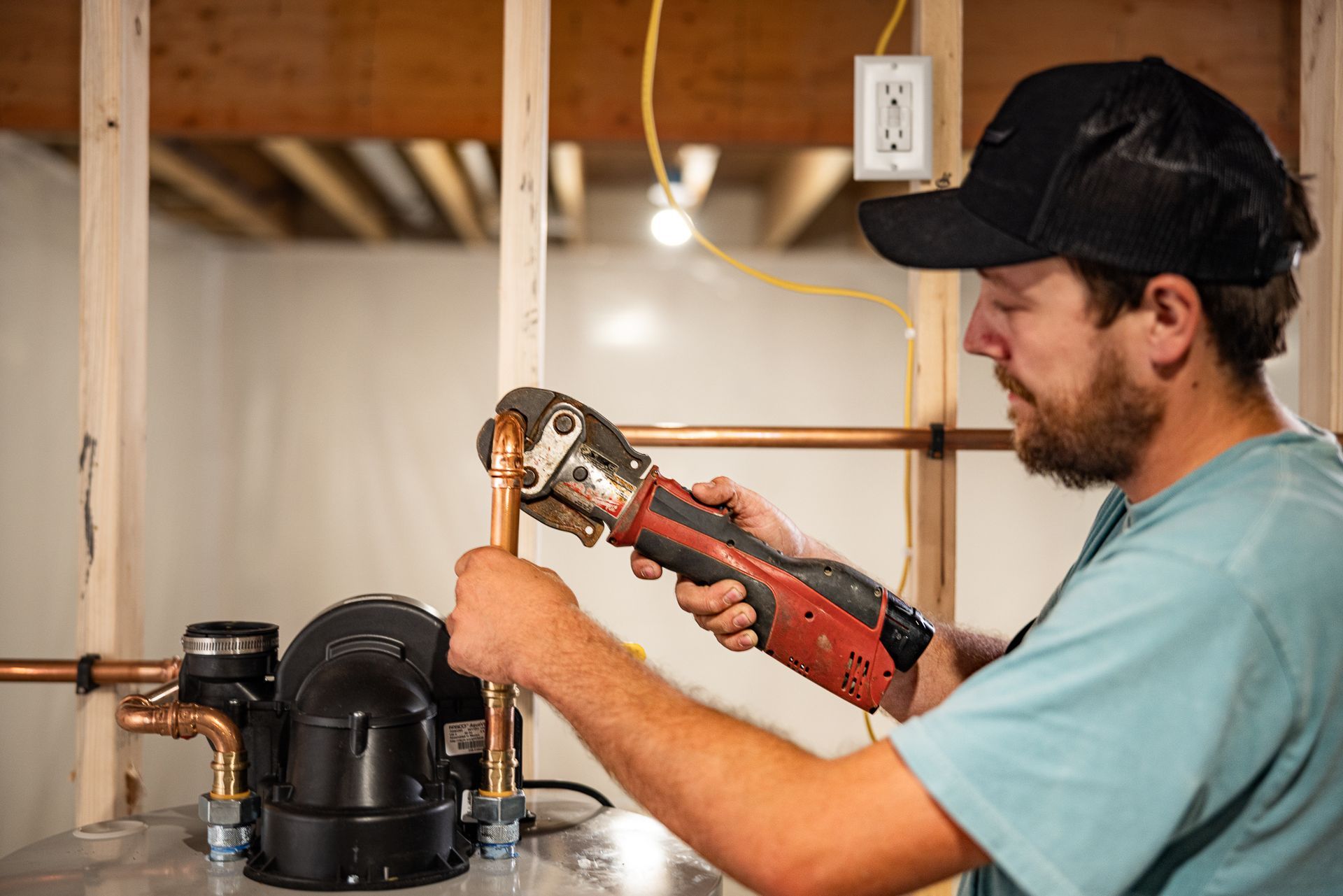 Man in cap and work clothes inspects pipes near the floor of a basement.