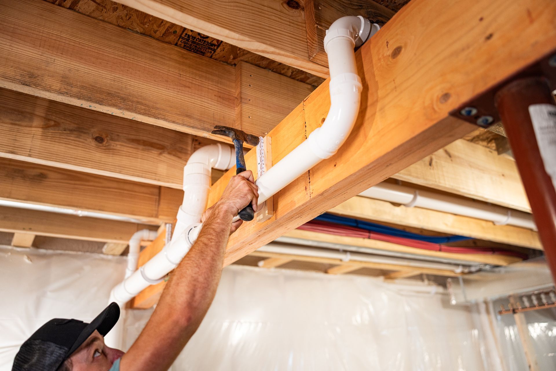 Man hammers pipe to wooden beam in basement; white pipes, wooden beams, and a black cap.