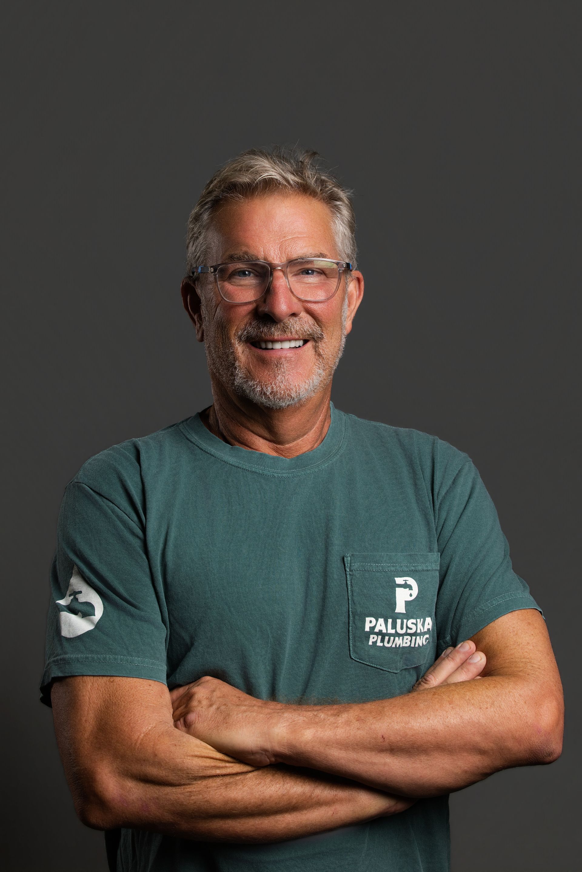 Smiling man with glasses and crossed arms, wearing a teal T-shirt against a dark gray background.