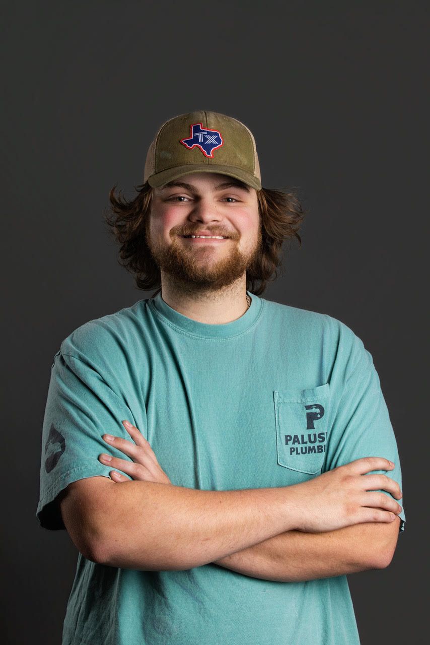 Smiling person in a tan cap and teal T-shirt with arms crossed against a dark background