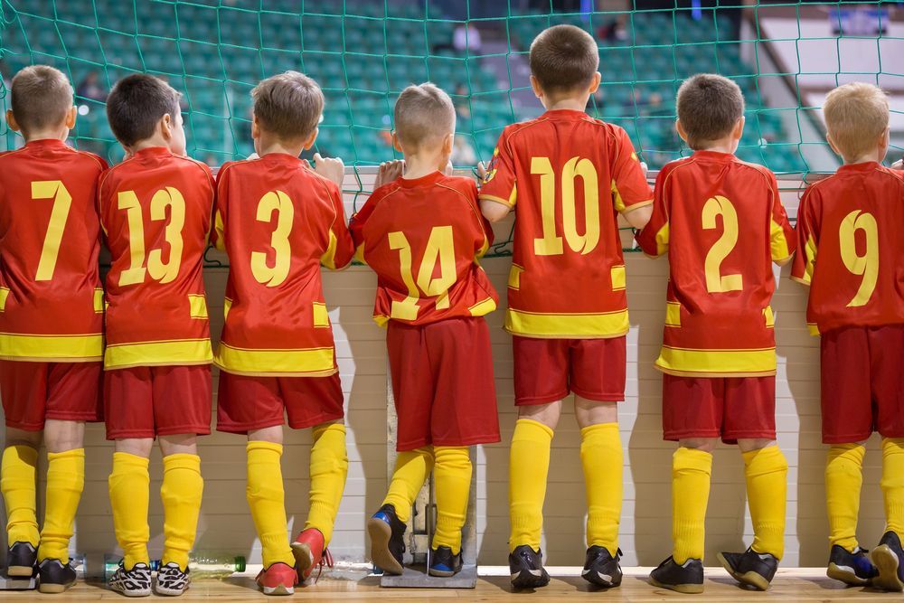 Siete niños con uniformes de fútbol rojos y amarillos idénticos están de pie en fila, de espaldas a la cámara, mirando un estadio.