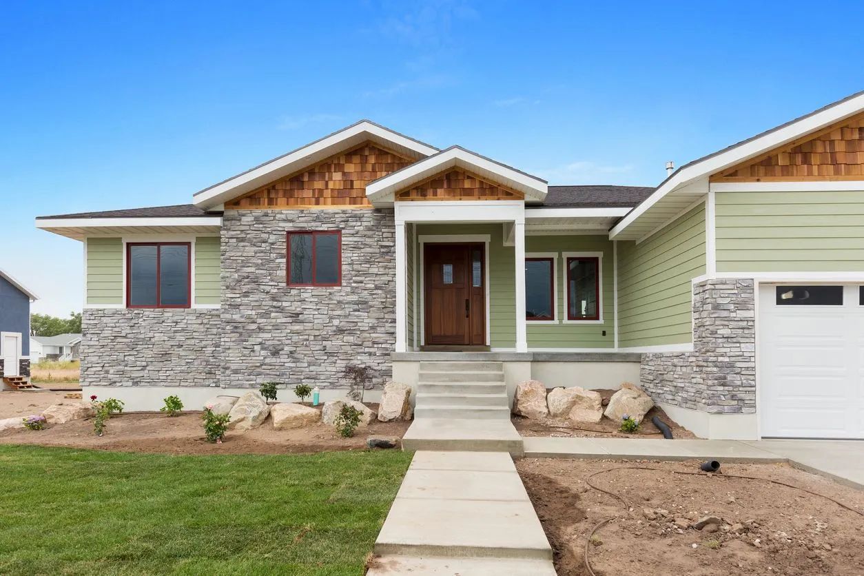 Ranch-style home with stone and green siding, brown door, and a concrete walkway on a sunny day.