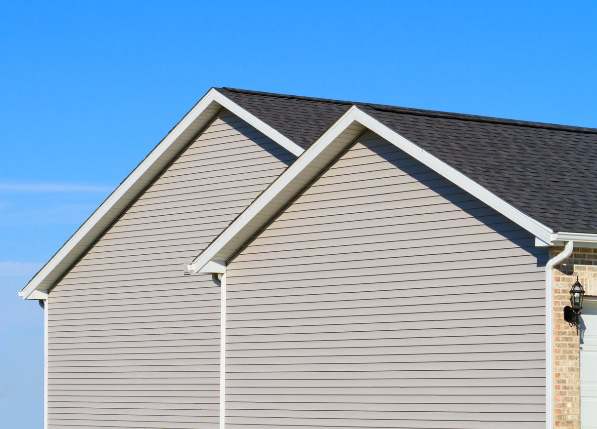 Two gabled roofs with dark shingles sit atop light-colored horizontal siding against a clear blue sky.