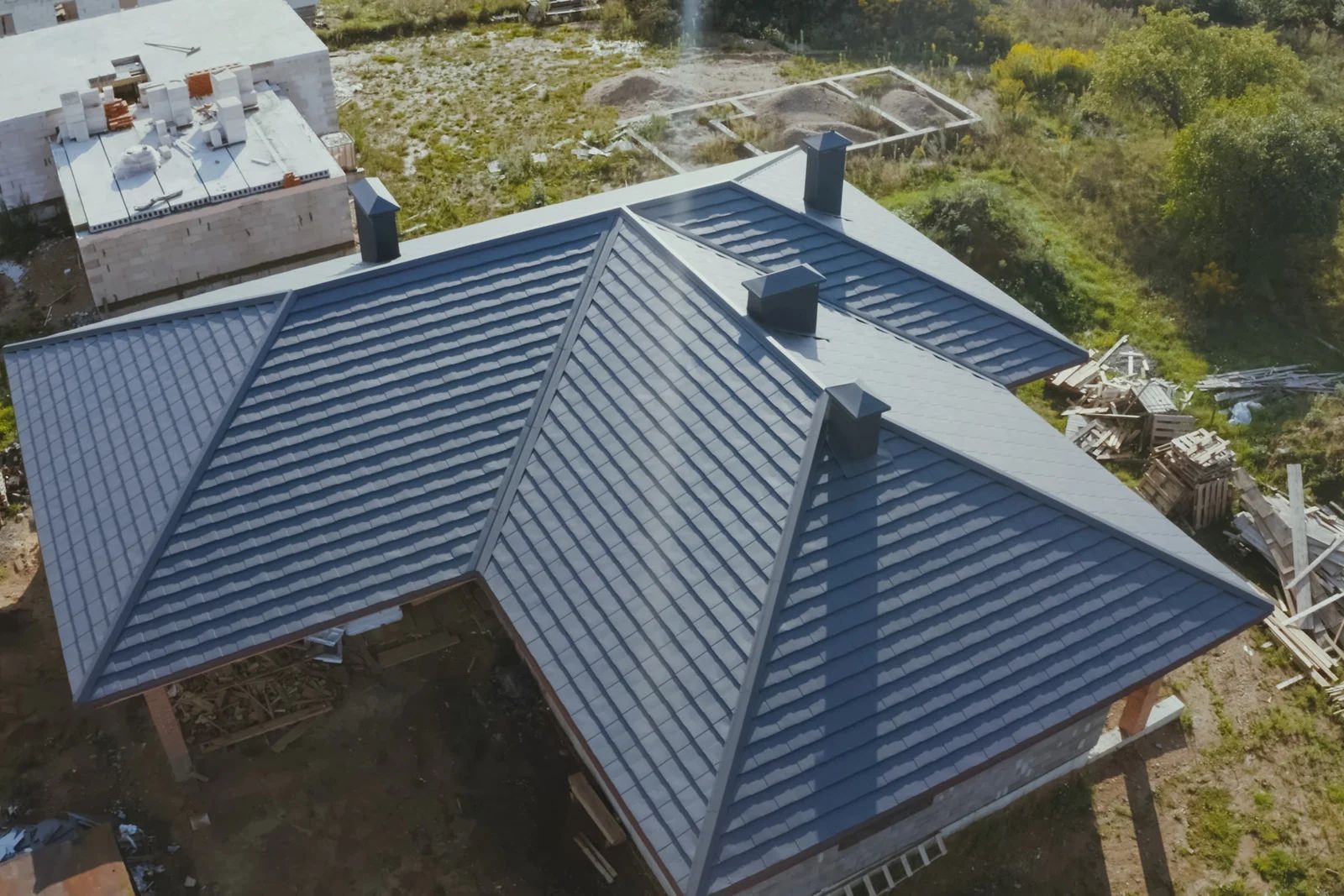 An aerial view of a dark grey metal hip roof on a building under construction, featuring multiple chimneys.