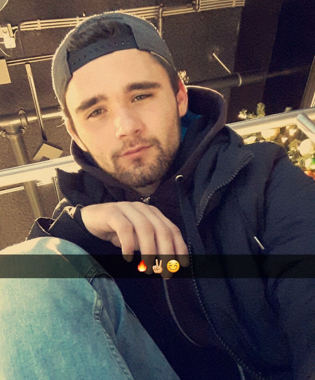 Een man met een baard en een baseballpet, gekleed in een zwarte jas, maakt een selfie.