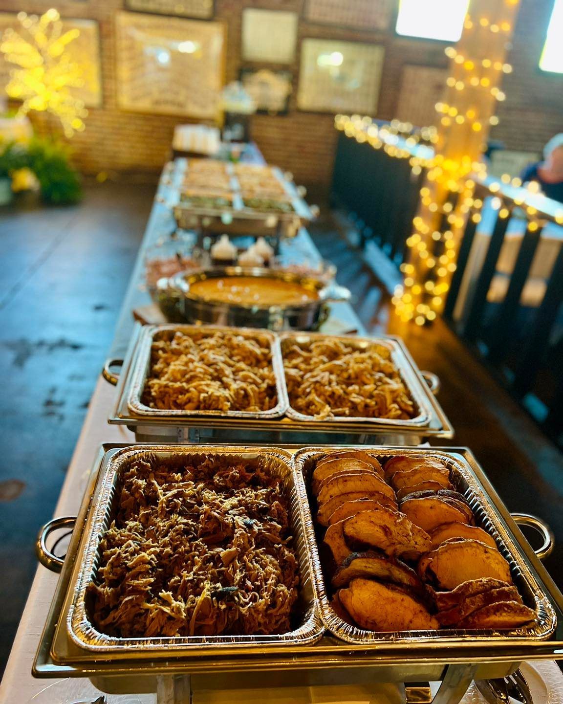 Buffet table with trays of assorted hot dishes at an outdoor evening event, lit by string lights