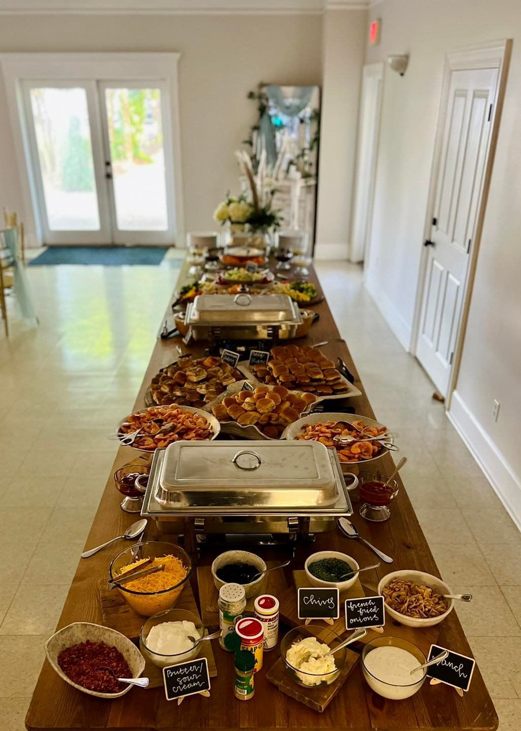 Buffet table with various dishes and condiments in a bright hallway leading to glass doors
