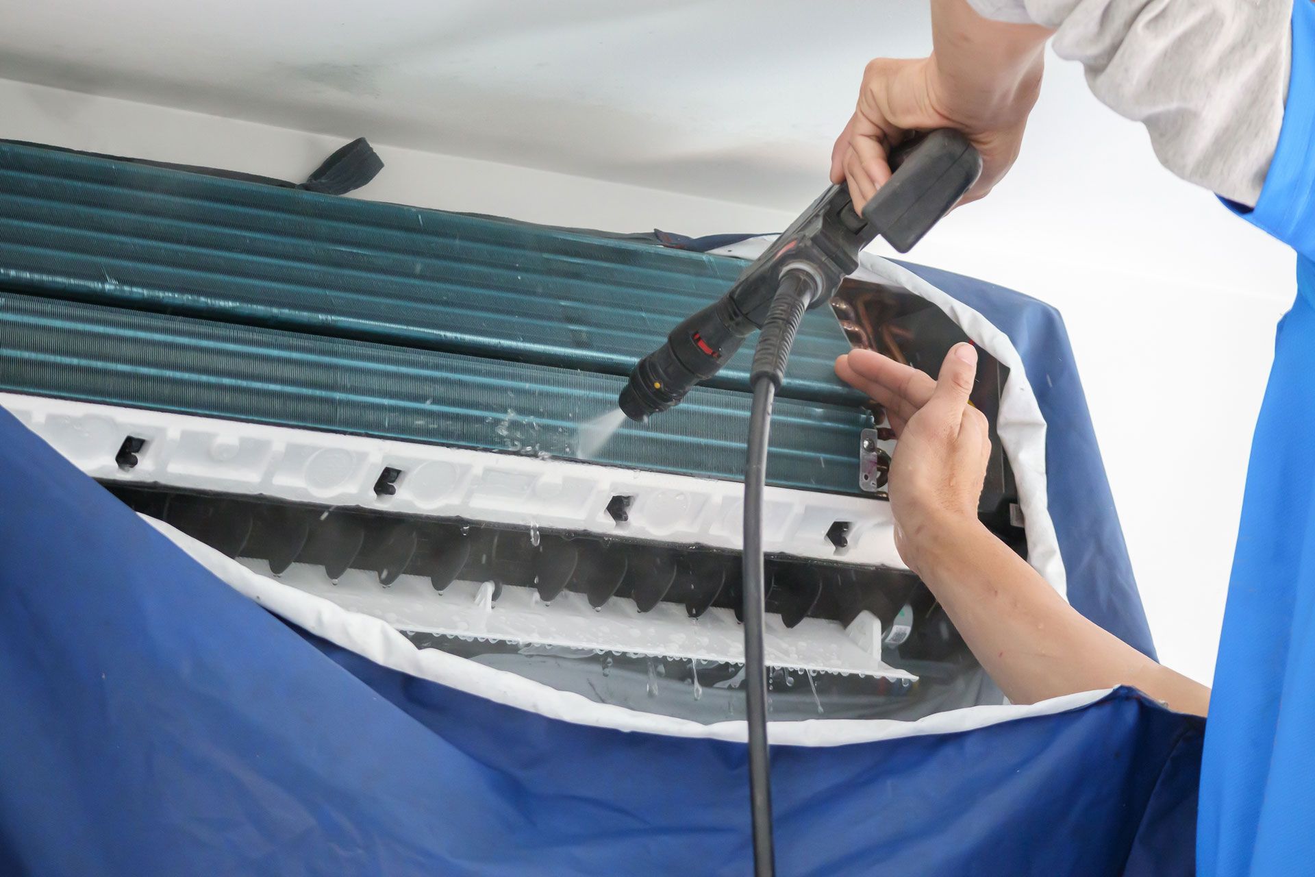 A person uses a pressure washer to clean the internal cooling fins of a wall-mounted air conditioner.