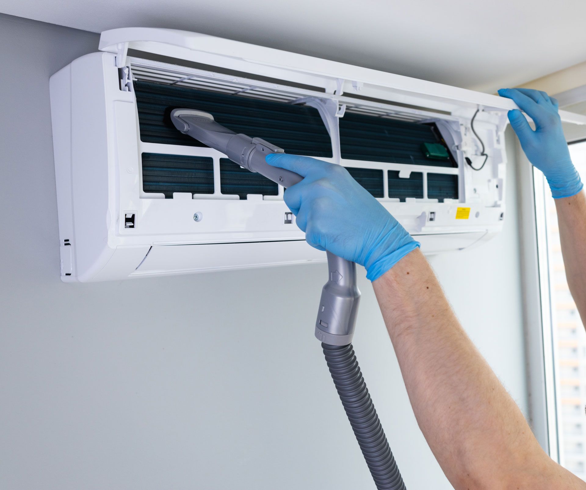 A person wearing blue gloves uses a vacuum cleaner nozzle to clean the dust filters of a white wall-mounted air conditioner.