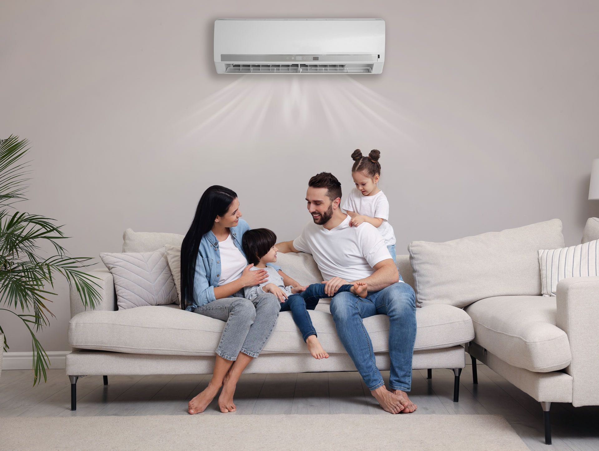 A family sits on a sofa under a wall-mounted air conditioner blowing cool air in a bright, modern living room.
