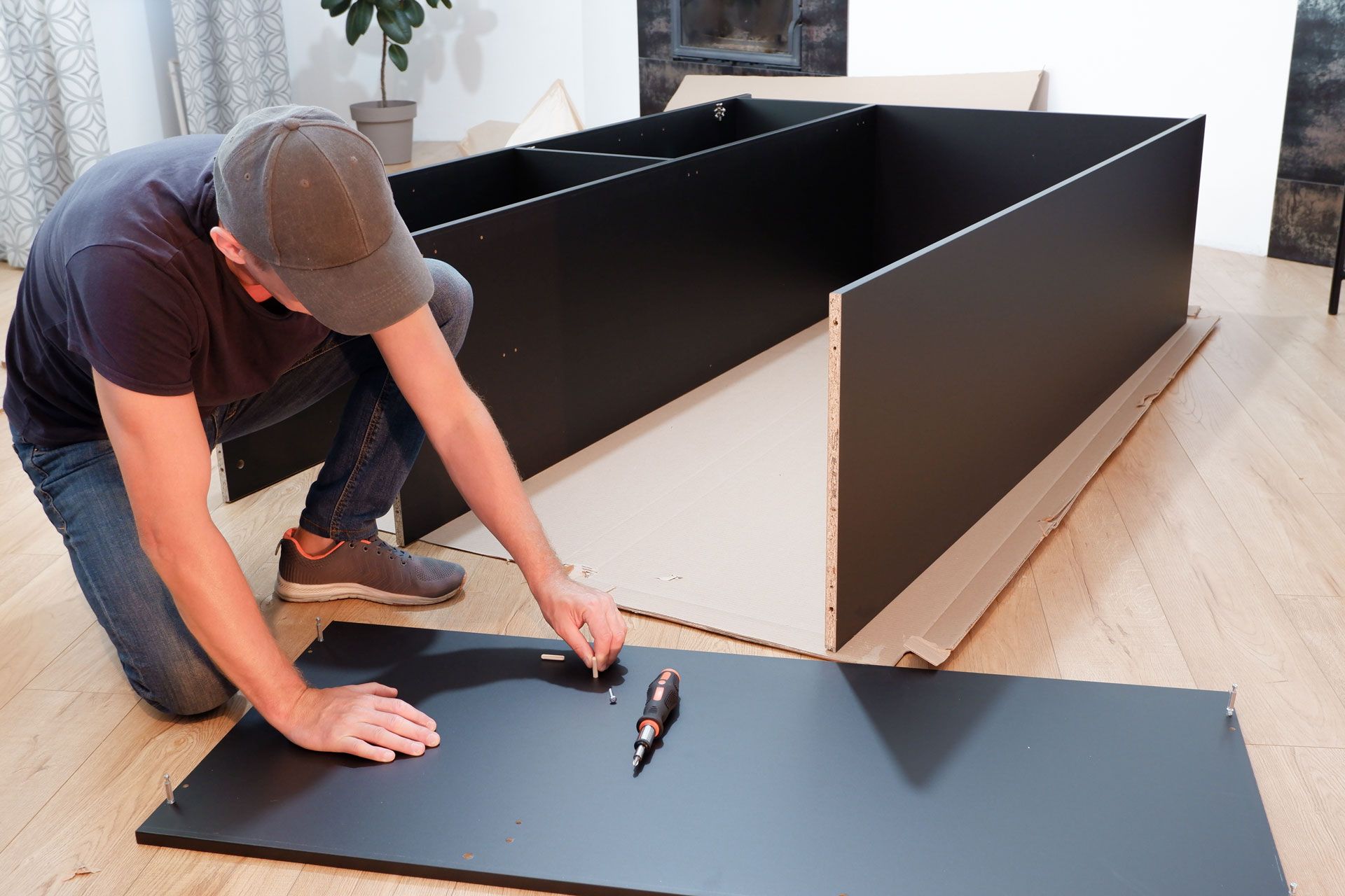 A person wearing a cap kneels on a wooden floor while assembling black furniture panels with a screwdriver.