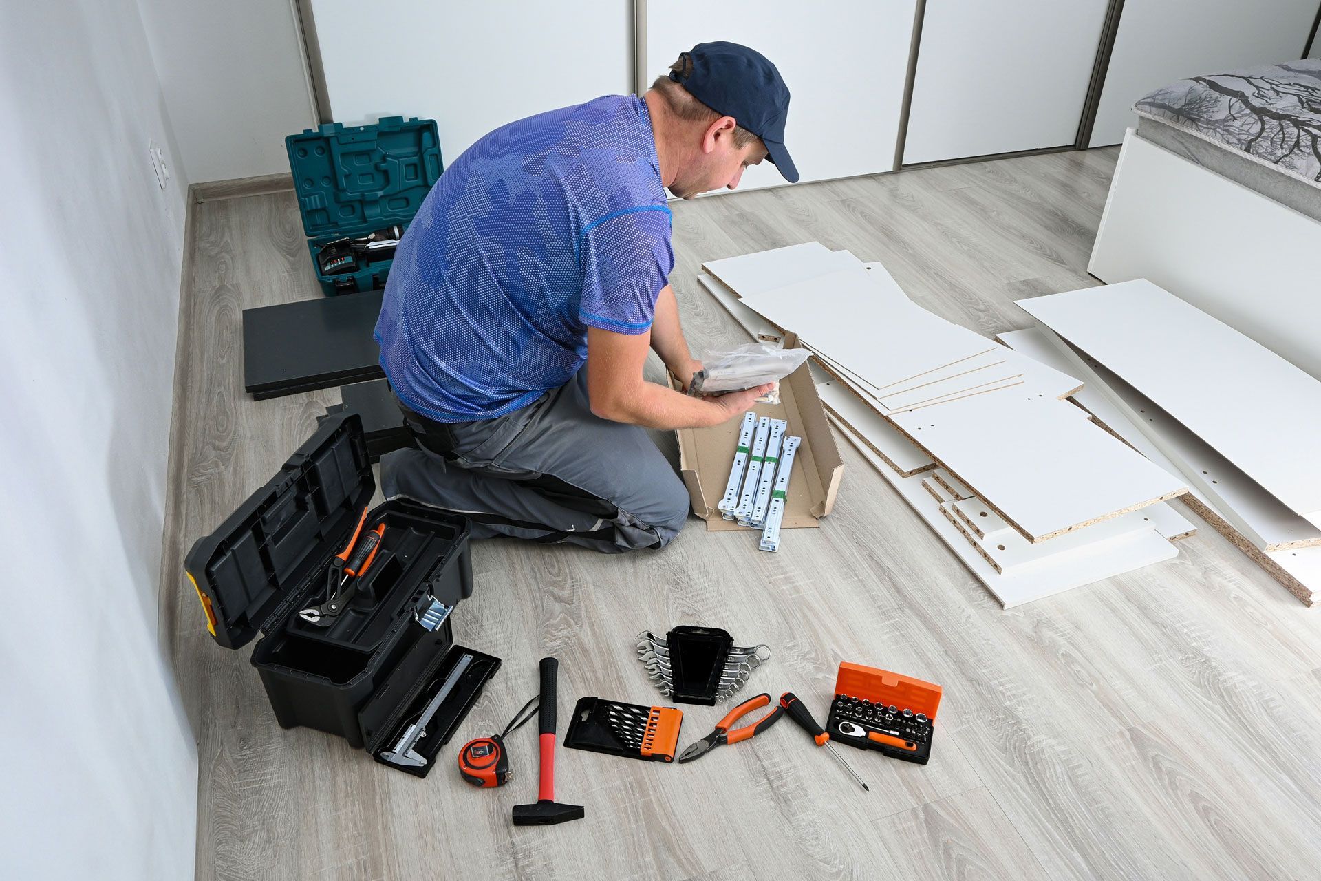 A person in a blue shirt kneels on a light floor, assembling white furniture pieces surrounded by tools in a bright room.