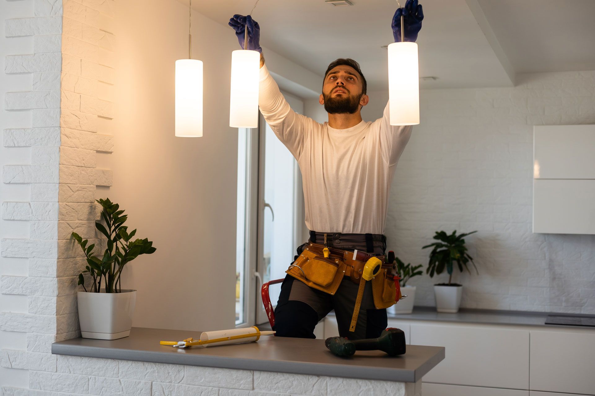 A worker wearing a tool belt installs pendant lights in a kitchen.