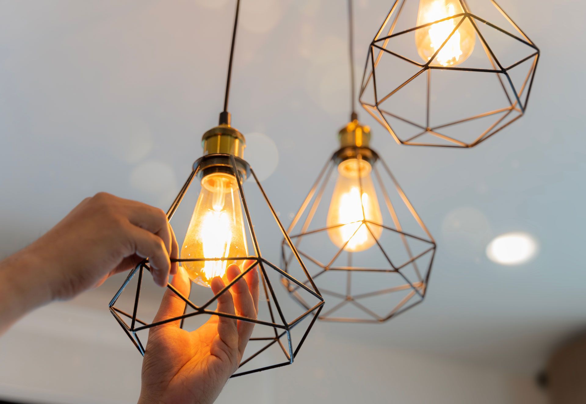 A pair of hands adjusting a glowing Edison-style lightbulb inside a geometric wire pendant lamp fixture.