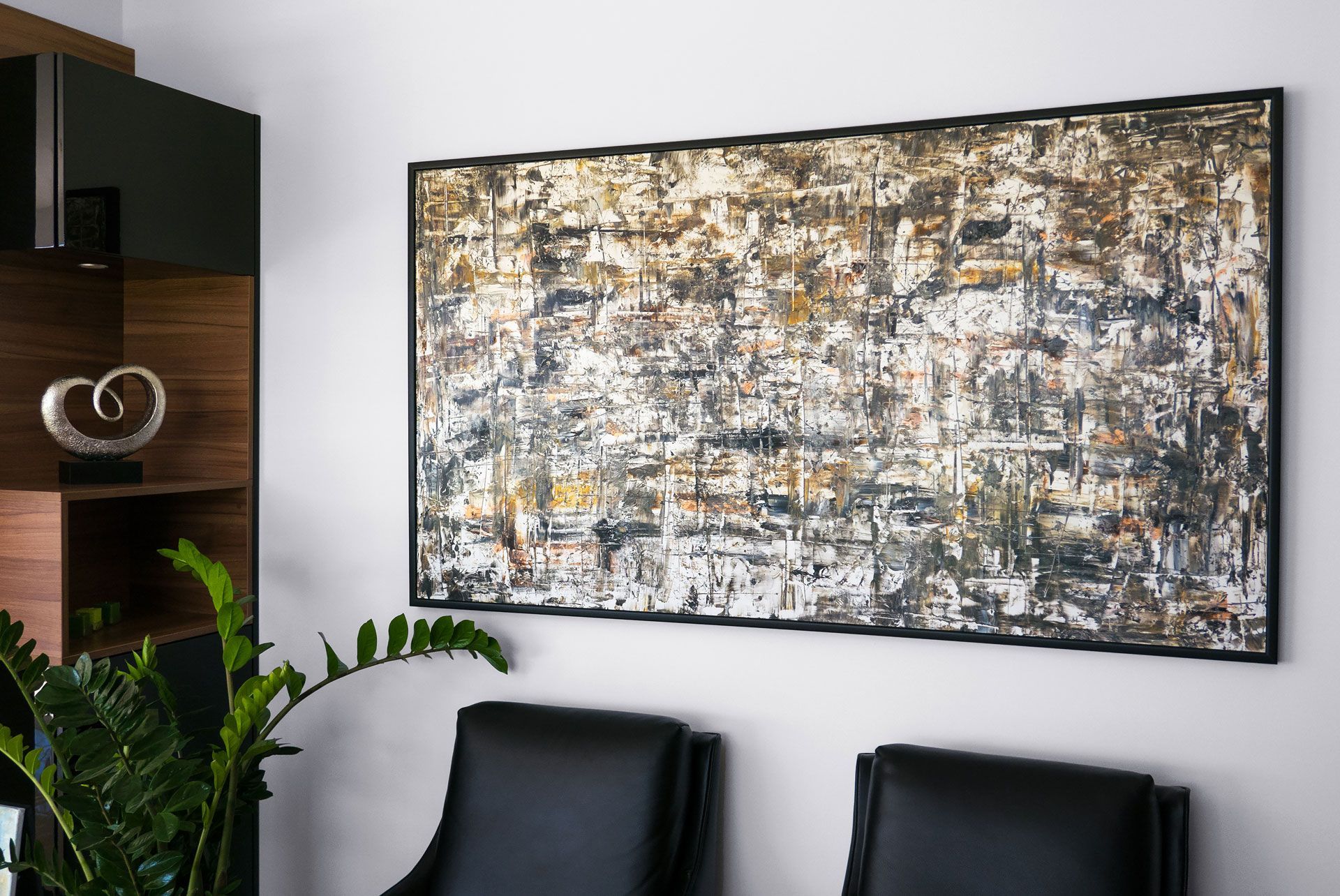 A large abstract painting with black, white, and gold tones hangs on a white wall above two black chairs and a plant.