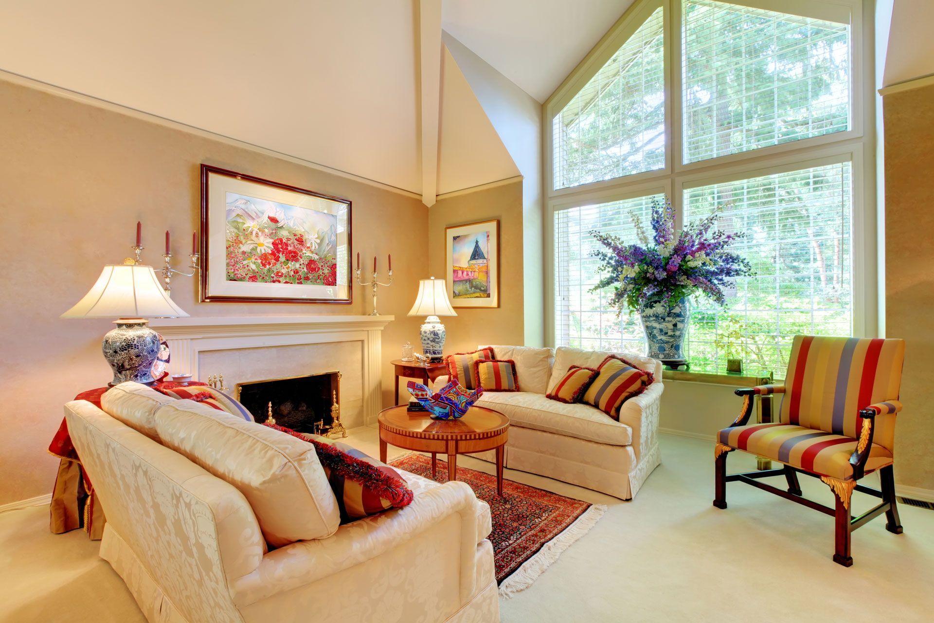 A sunlit living room with a fireplace, cream-colored sofas, a striped armchair, and large windows with garden views.
