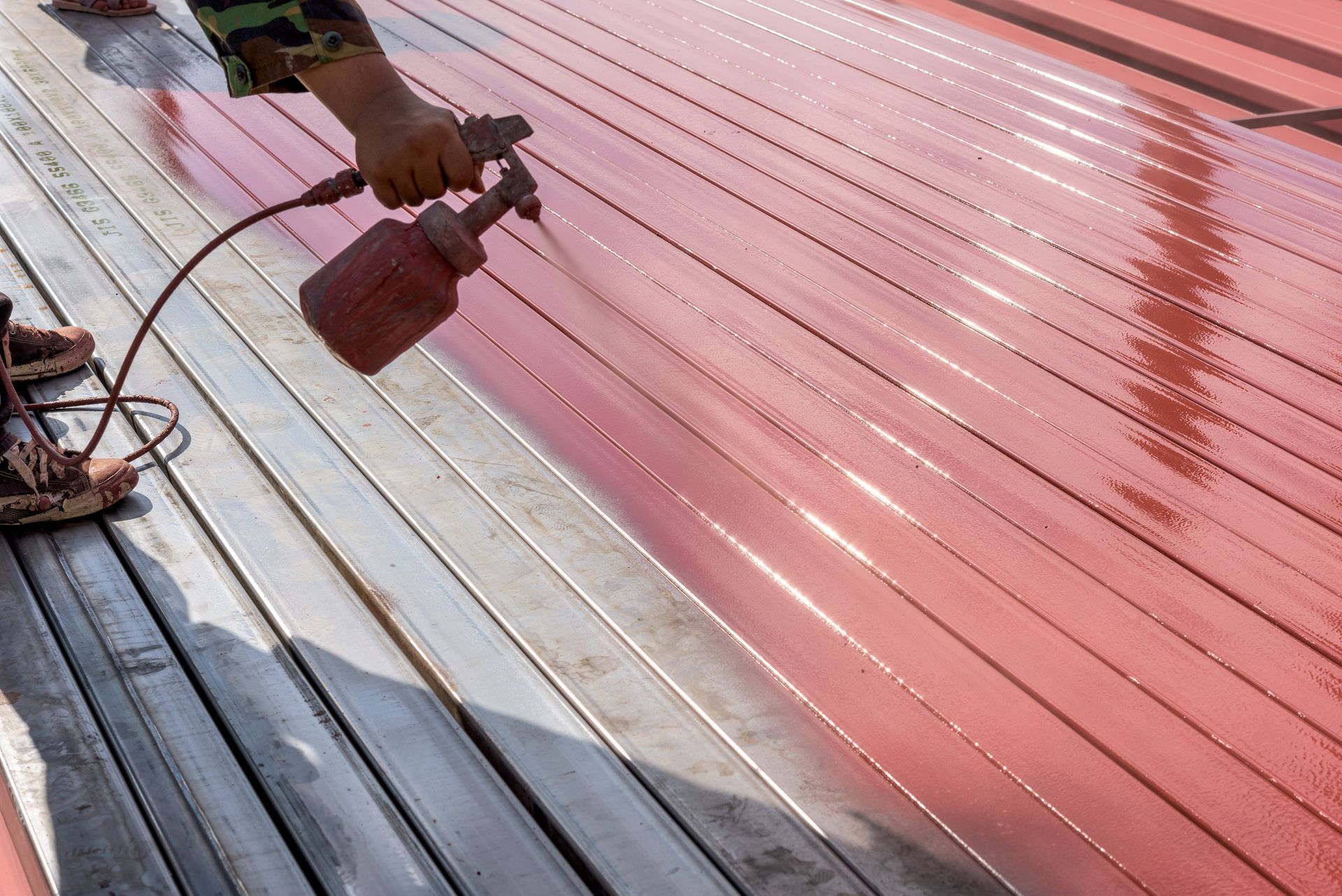 Metal roof of a building being painted with a vibrant red color using a spray technique.