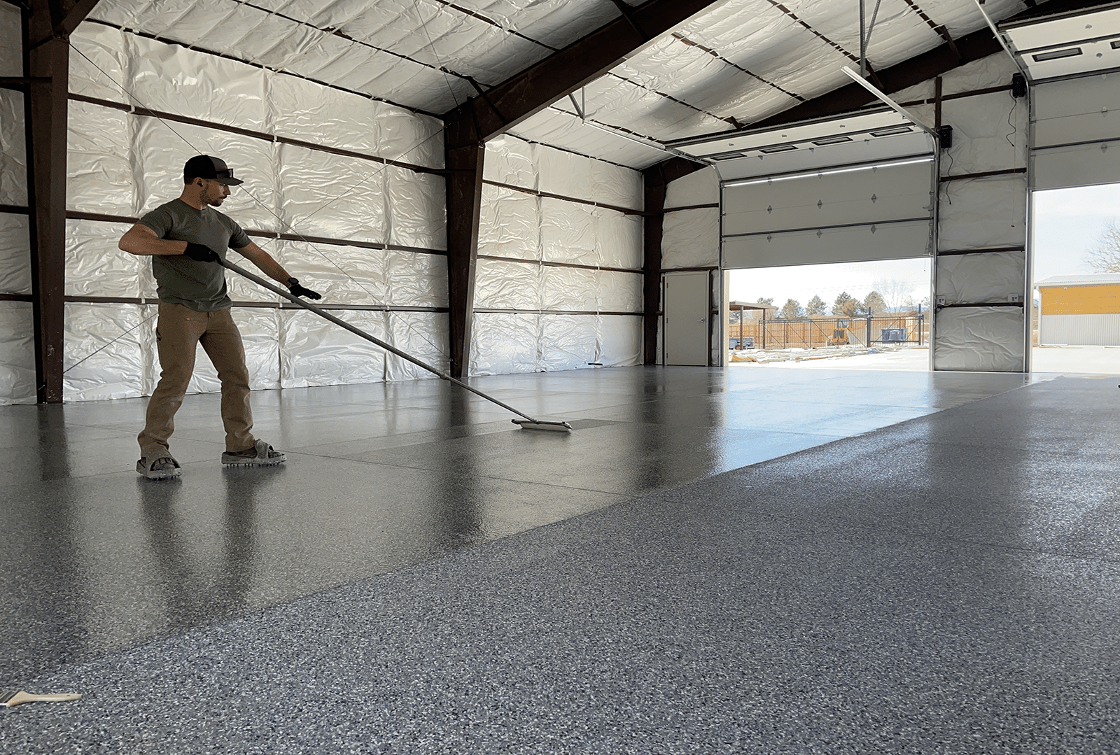 Man applying epoxy coating to a garage floor. The floor is shiny gray; building has an open door.