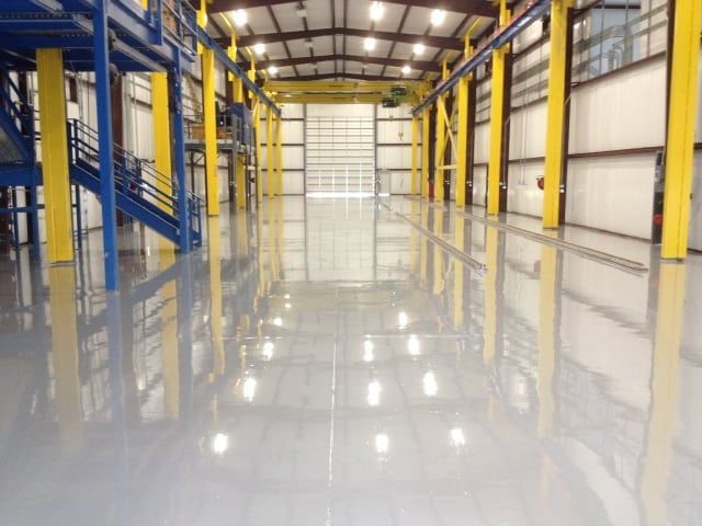 Empty industrial warehouse with reflective gray epoxy floor, yellow support beams, and overhead lights.