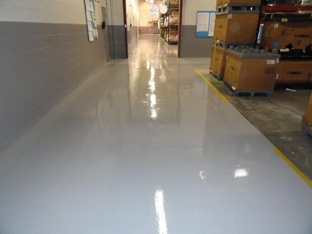 Shiny gray epoxy floor in a warehouse aisle. Yellow line marks edge near stacked boxes.