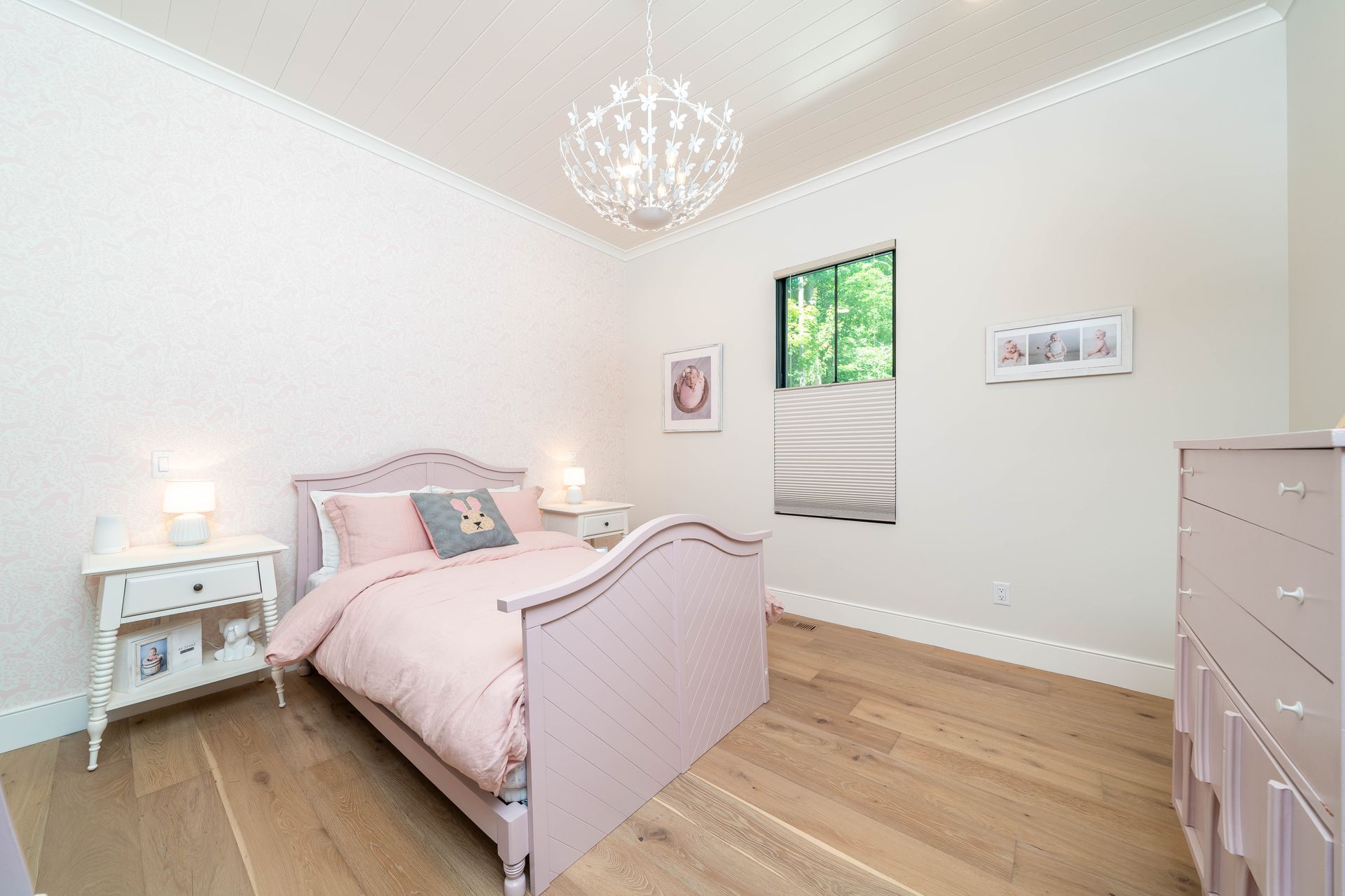 A bedroom with a pink bed , dresser , nightstand and window.
