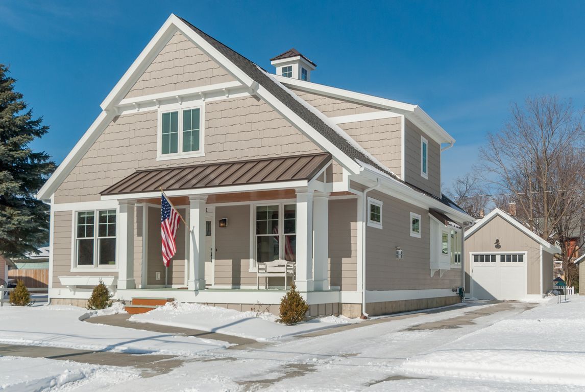 Beige house with a porch and detached garage, covered in snow on a sunny day.
