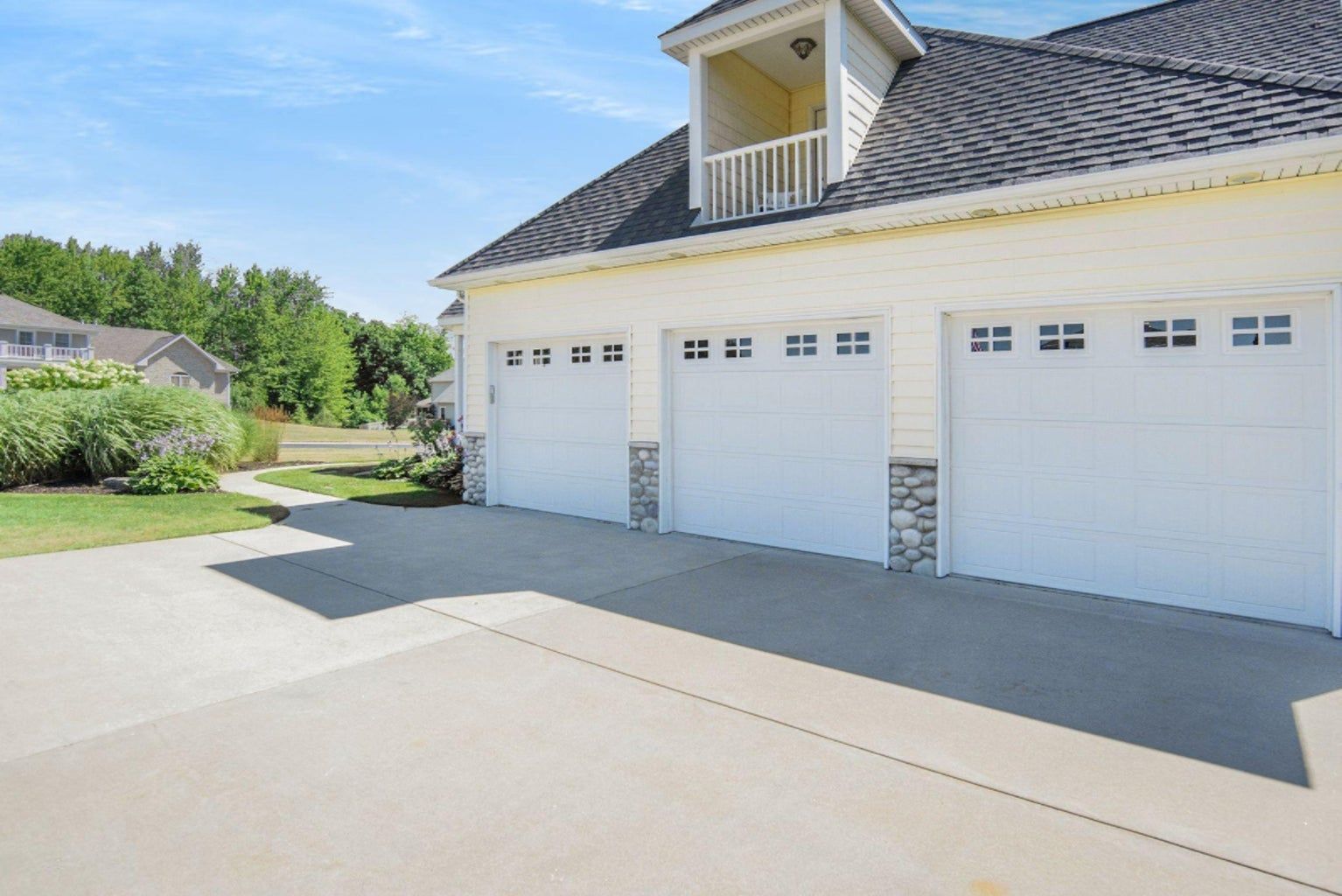 Three-car garage with white doors, light yellow siding, stone accents, and a concrete driveway on a sunny day.