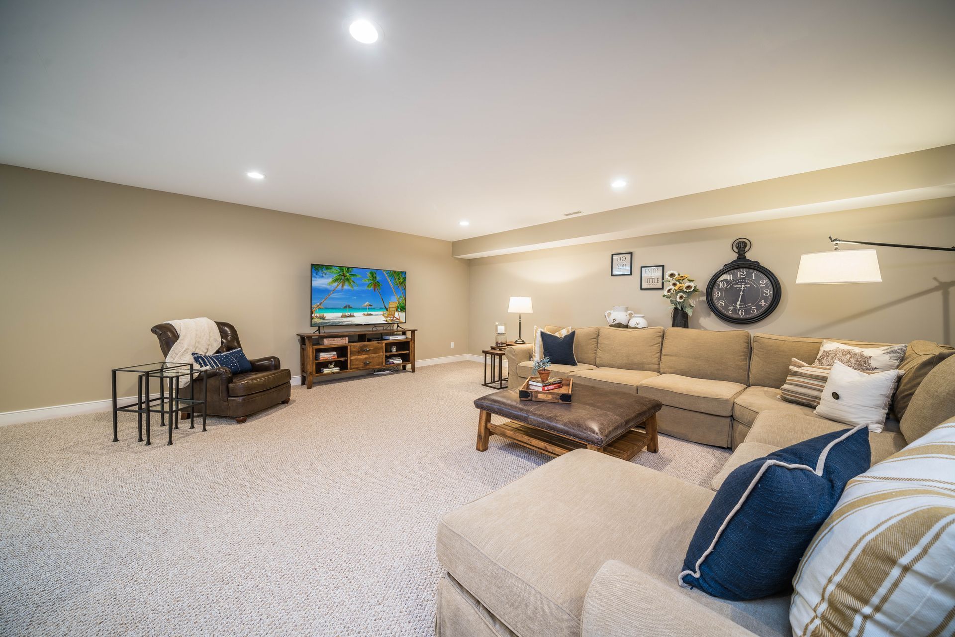 Spacious living room with sectional sofa, TV, armchair, coffee table, and carpet. Beige walls and ceiling with recessed lighting.