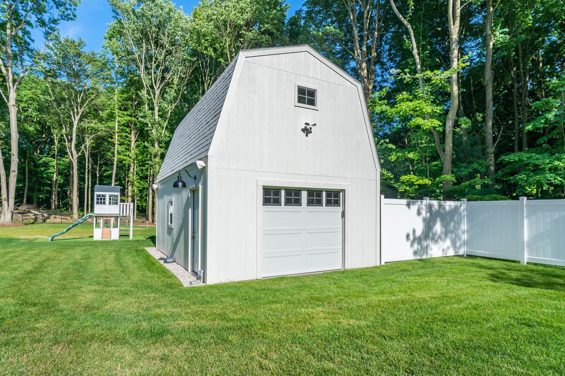 White barn-style garage with a closed door, surrounded by green grass and a white fence in a wooded area.