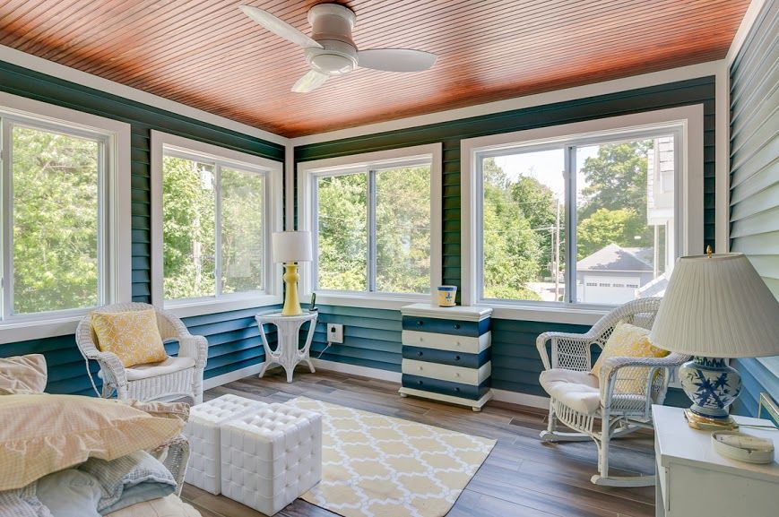 Sunlit screened porch with blue-and-white seating, striped cabinets, and large windows facing greenery