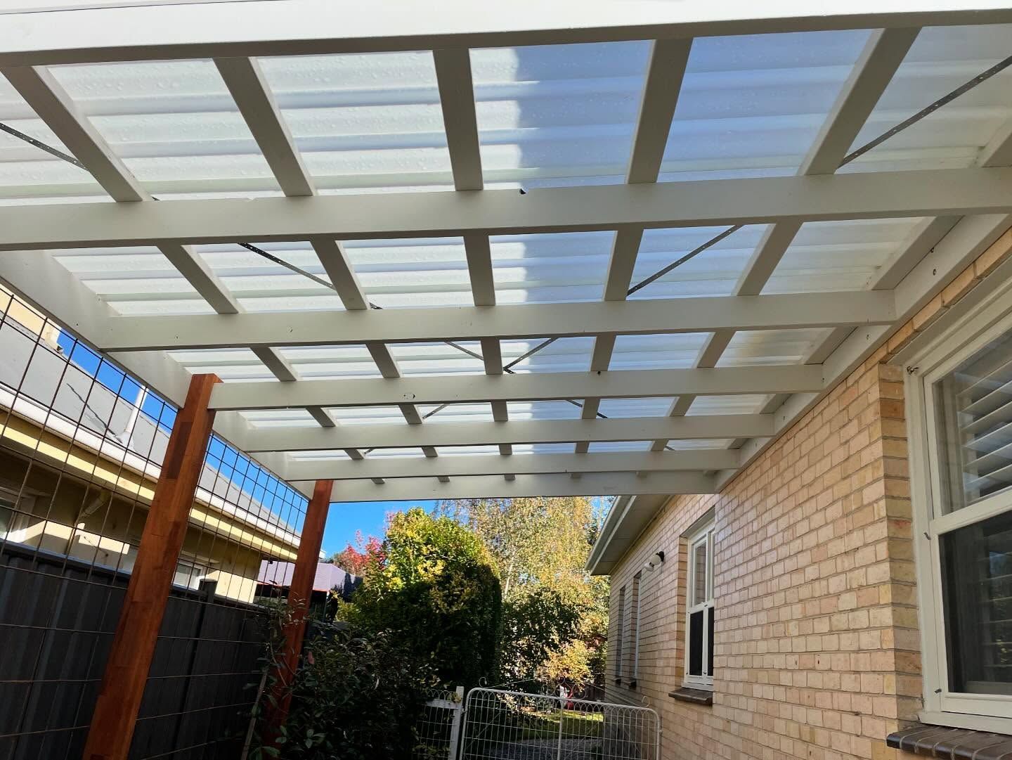 White-framed pergola with clear roofing over a brick building's side, with sunlight filtering through the roof.