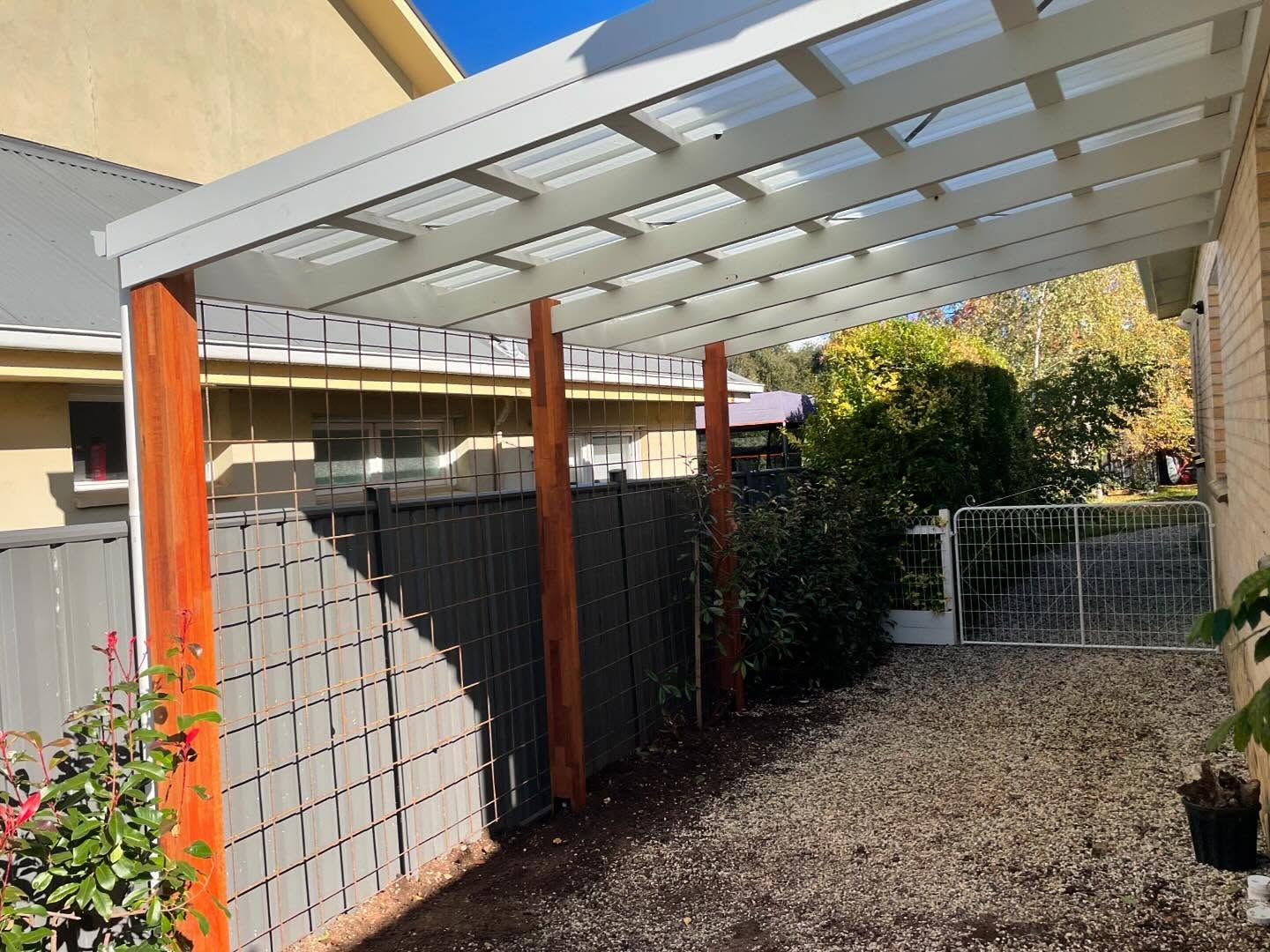 Wooden carport with clear roof over gravel path beside a gray fence and bushes.