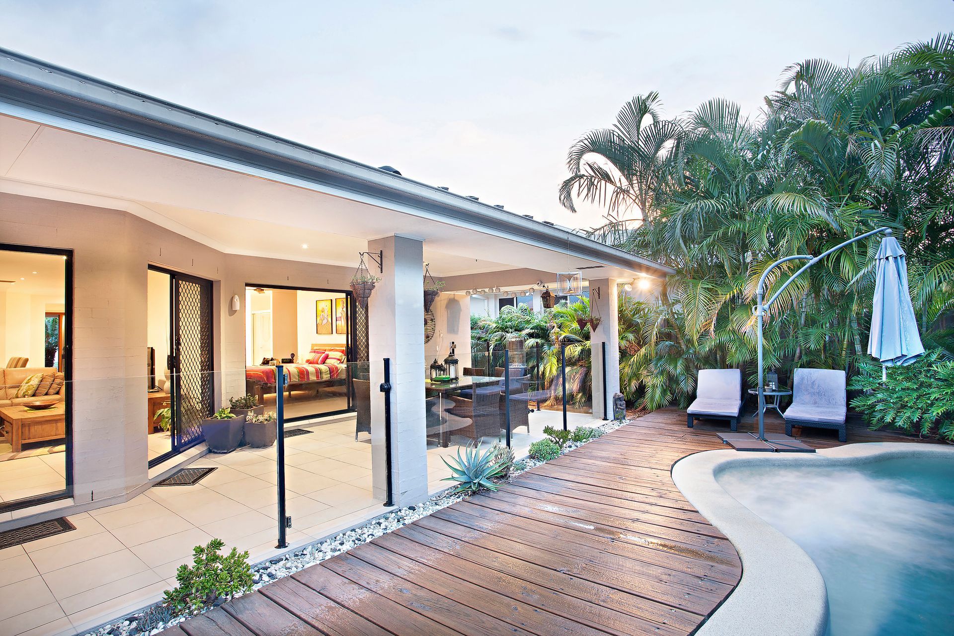 Patio with pool and covered dining area at dusk. Brown wood deck, plants, and outdoor furniture.