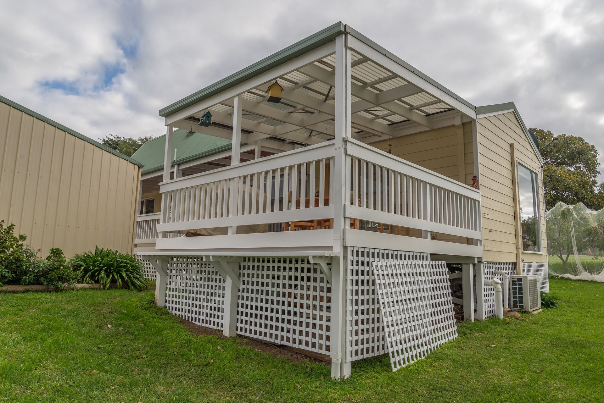 White deck with lattice skirting, green roof, overlooking a grassy area under a cloudy sky.