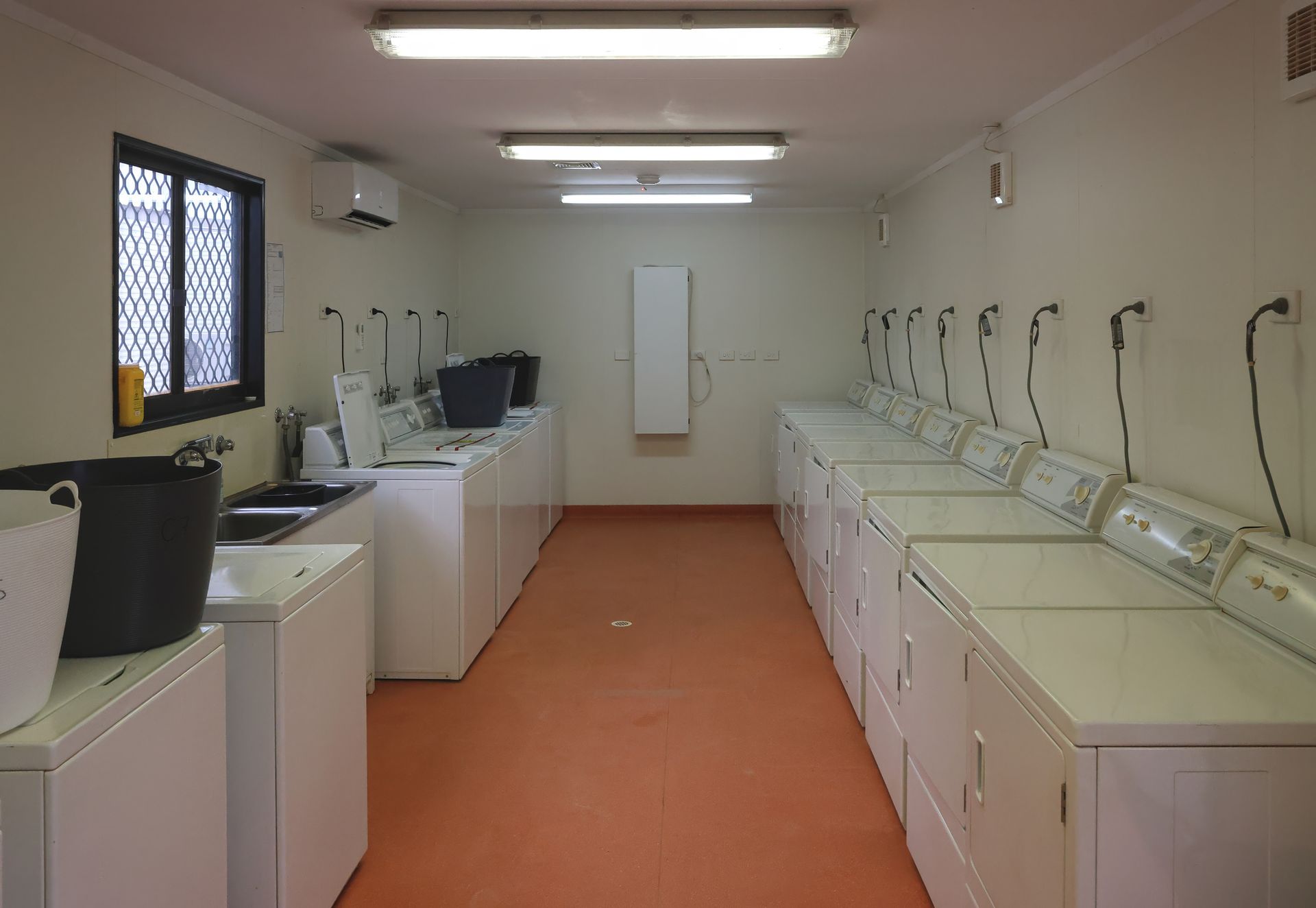 Laundry room with rows of white washers and dryers. Orange floor, white walls, and fluorescent lighting.
