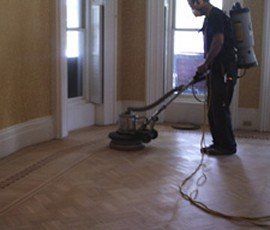 Man sanding hardwood floor in a room with tall windows and light-colored walls, using a large sander.