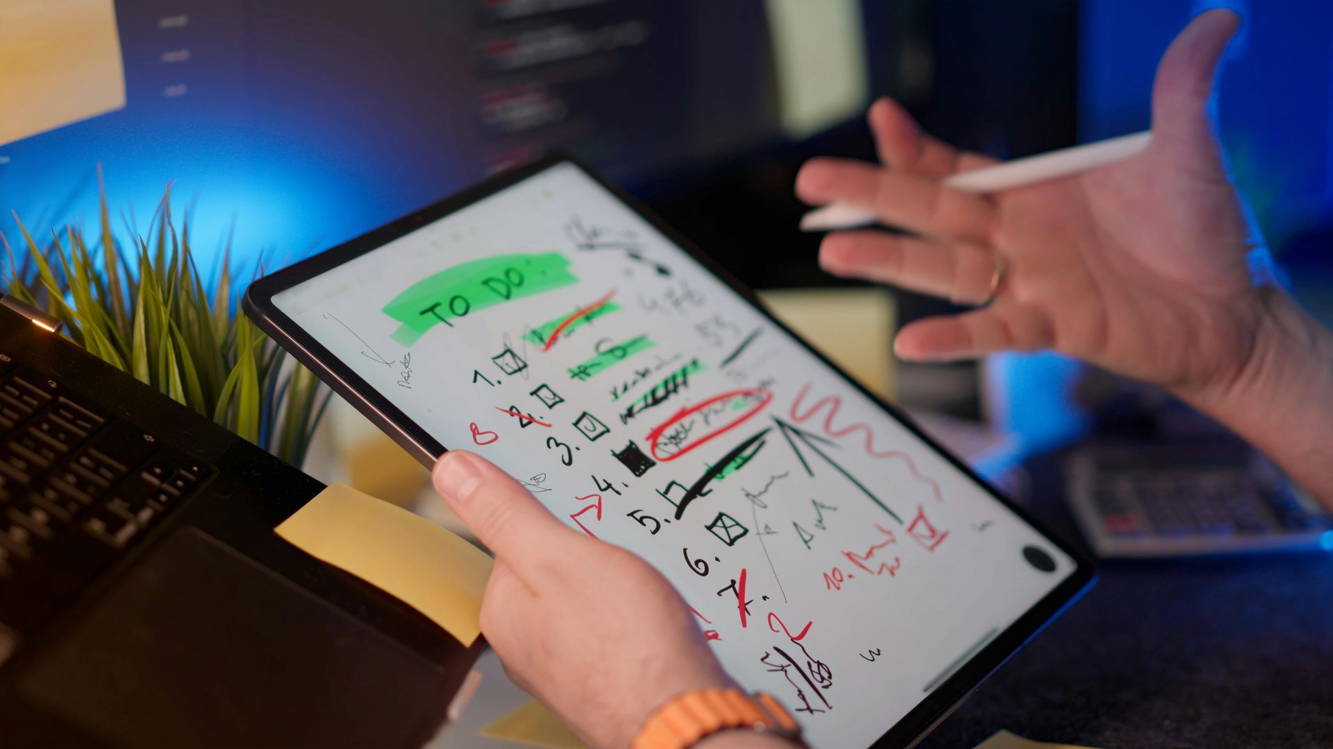 Hands holding a tablet with a to-do checklist and red markings on a desk, lit by blue and warm light