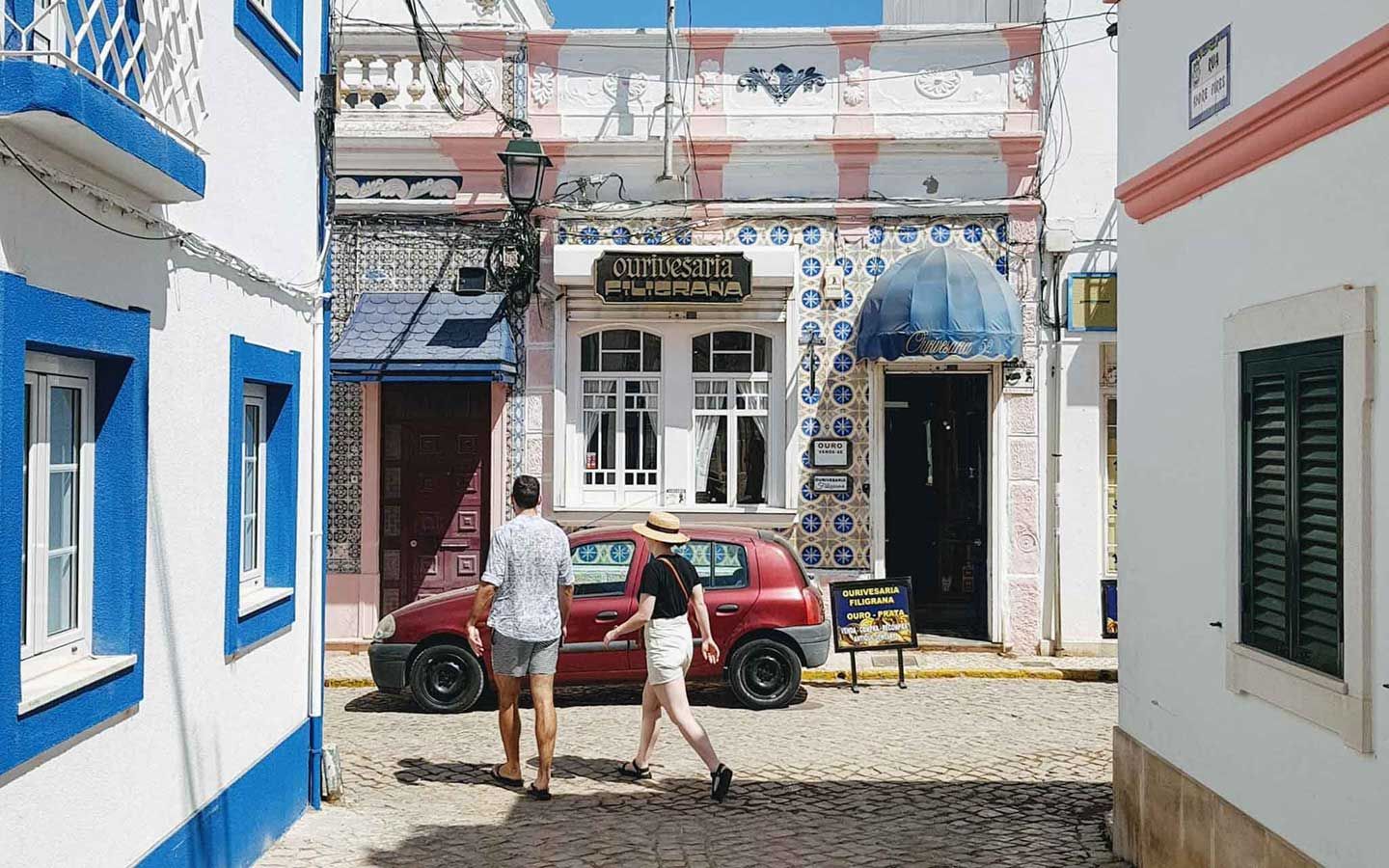 A man and woman are walking down a cobblestone street next to a red car.