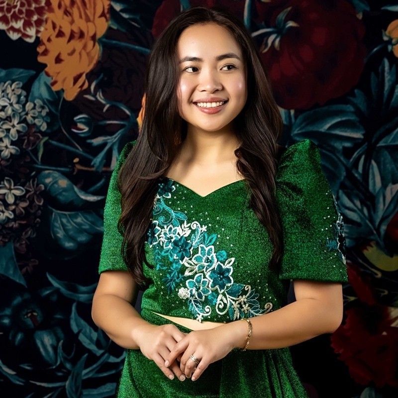 A woman in a green dress is smiling in front of a floral background.